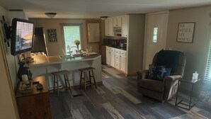 A modern kitchen with a central island, bar stools, and a dining area with a sofa and chair. - The Rustic Minnow: Walk to Lake, Enjoy Outdoor Amenities and Stunning Landscape  (Hulbert)