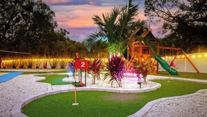 Um jardim bem cuidado com área de recreação, incluindo escorregador e estruturas para escalar, cercado por sebes podadas e palmeiras.