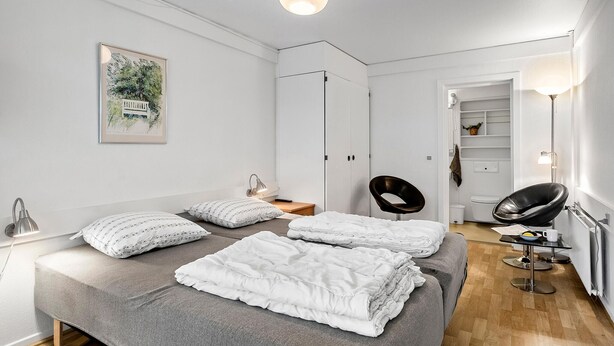 Ein modernes Schlafzimmer mit einem Bett, zwei Sesseln, einem Nachttisch, einer Lampe und einem Kleiderschrank.