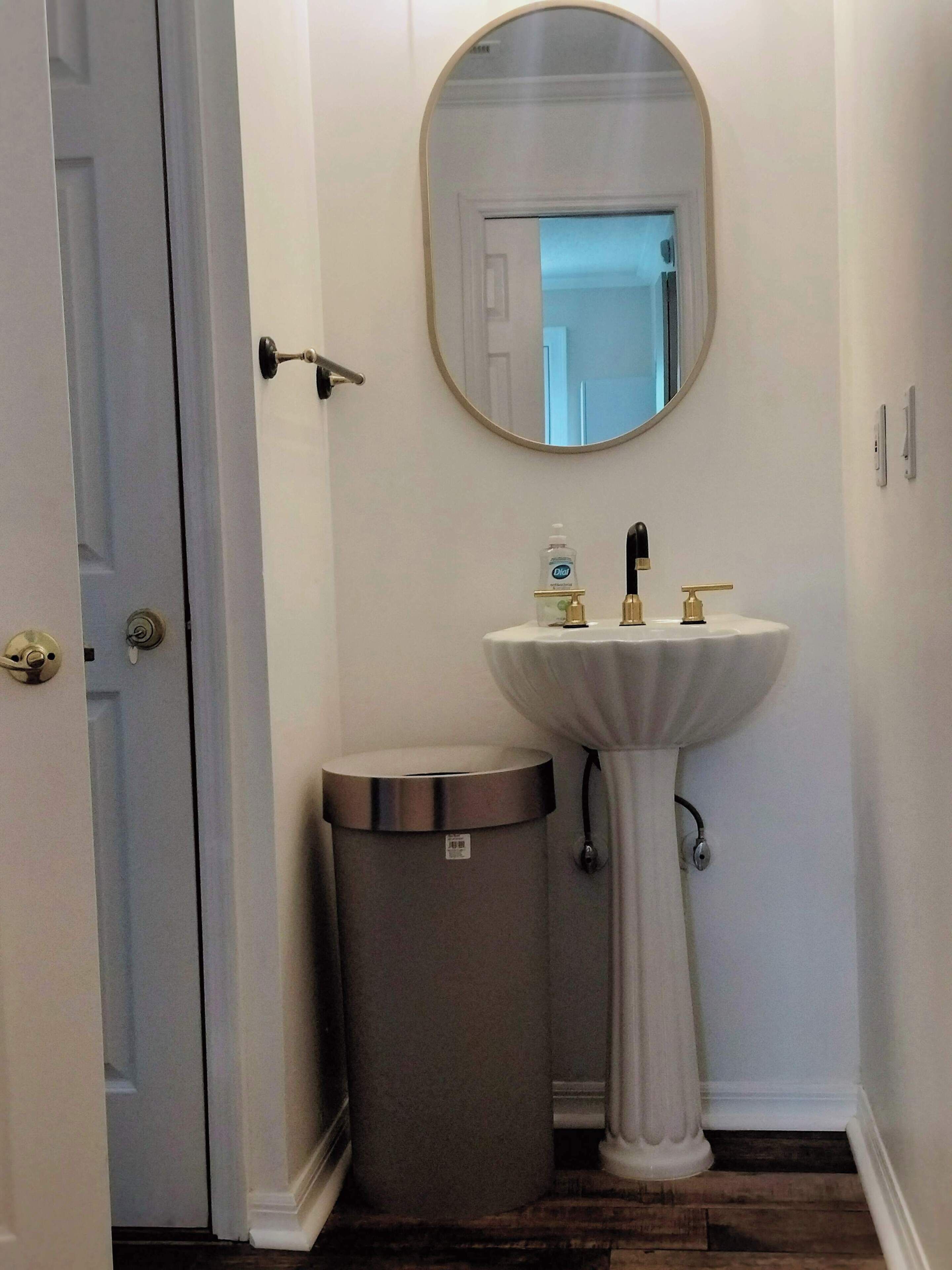 Ein Badezimmer mit einem Standwaschbecken, einem runden Spiegel, einem Mülleimer und einer Tür.