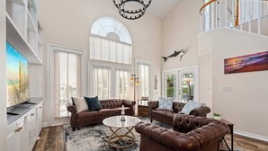 A spacious living room with a large window, a chandelier, and a staircase.
