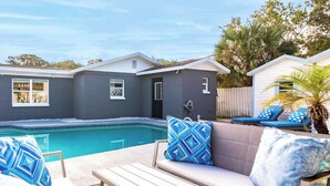 Ein Poolbereich mit Holzterrasse, einer beigen Couch mit gemusterten Kissen und einem Pool.