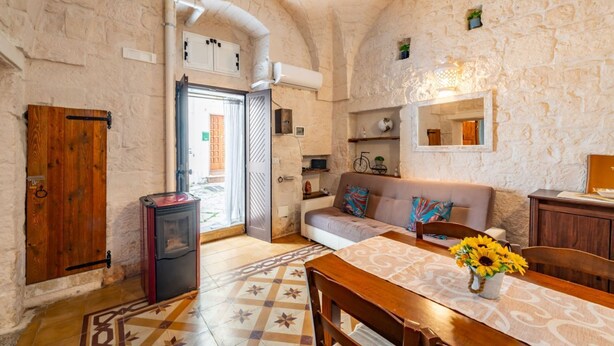 A rustic interior with a stone wall, a wooden door, a sofa, a table with a vase of sunflowers, and a small heater. - Antica Casa 1600 by Rentbeat (Ostuni)