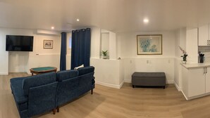 A modern living room with a blue sofa, a small table, and a TV. There is a kitchen with white cabinets and a microwave.