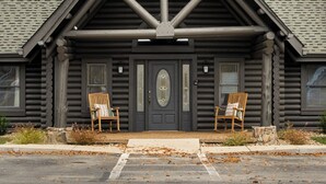A log cabin with a triangular roof, a central door, and two wooden chairs in front.