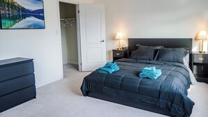 A bedroom with a dark blue bed, a white door, a coat rack, a dresser, and a nightstand with a lamp.