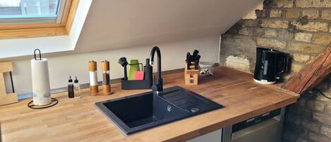 Una cocina con encimera de madera, un lavamanos negro y un lavavajillas. Hay una ventana sobre el lavamanos, una pared de ladrillo y una cafetera en la encimera.