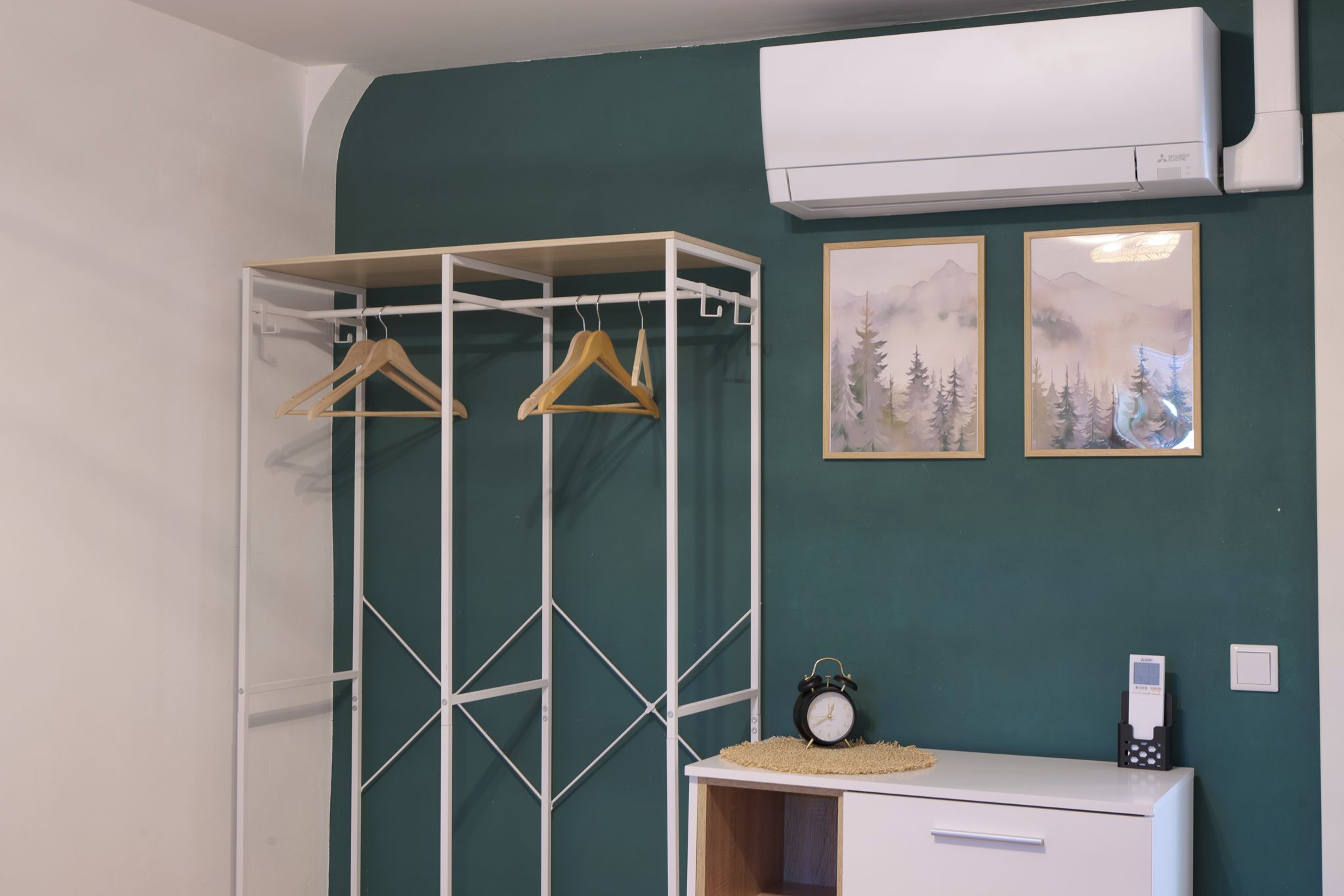 Ein Zimmer mit einem weißen Kleiderschrank, einem Kofferständer mit Holzaufhängern, einem kleinen Tisch mit einer Uhr und zwei gerahmten Bildern an der Wand.