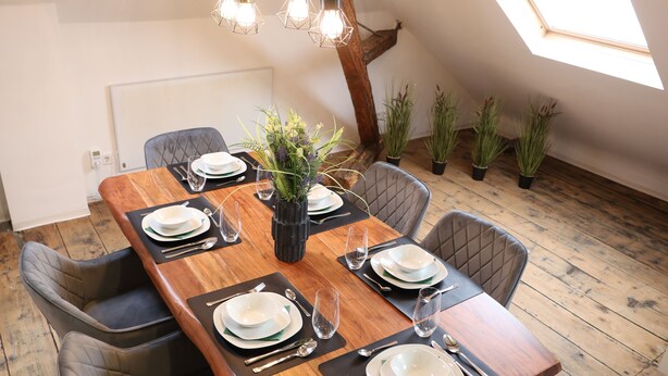 A dining table set for six with black placemats, white plates, and silverware. There are potted plants on the floor and a skylight above. - Charming half-timbered apartment with modern comfort, 110 sqm (Eltville am Rhein)