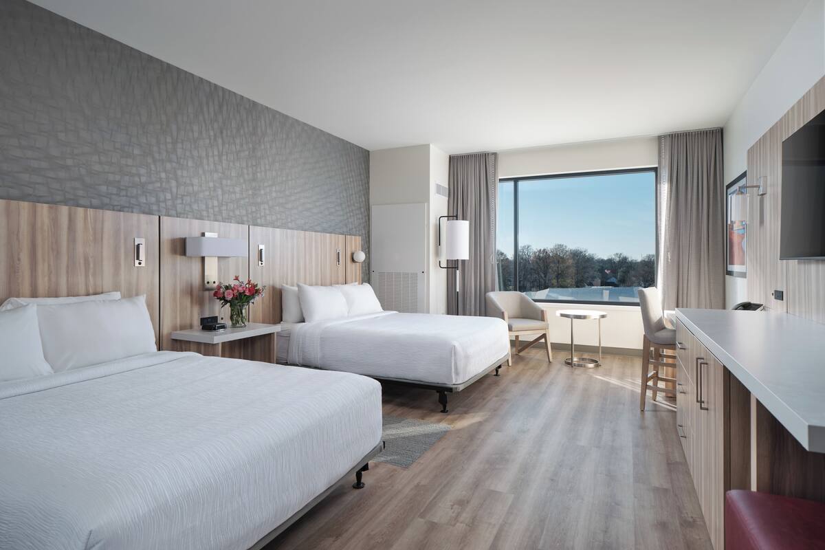 Habitación de hotel con dos camas, un escritorio, una silla, un televisor y un ventanal con vistas.