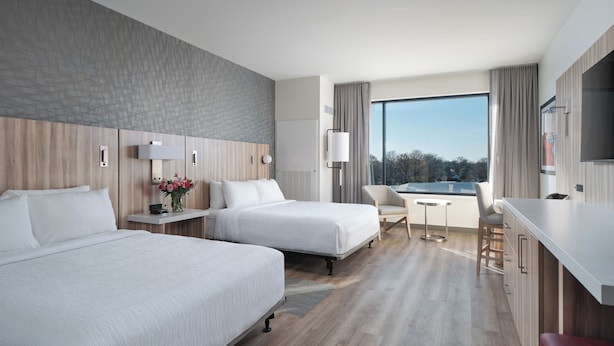 A hotel room with two beds, a desk, a chair, a TV, and a large window with a view.