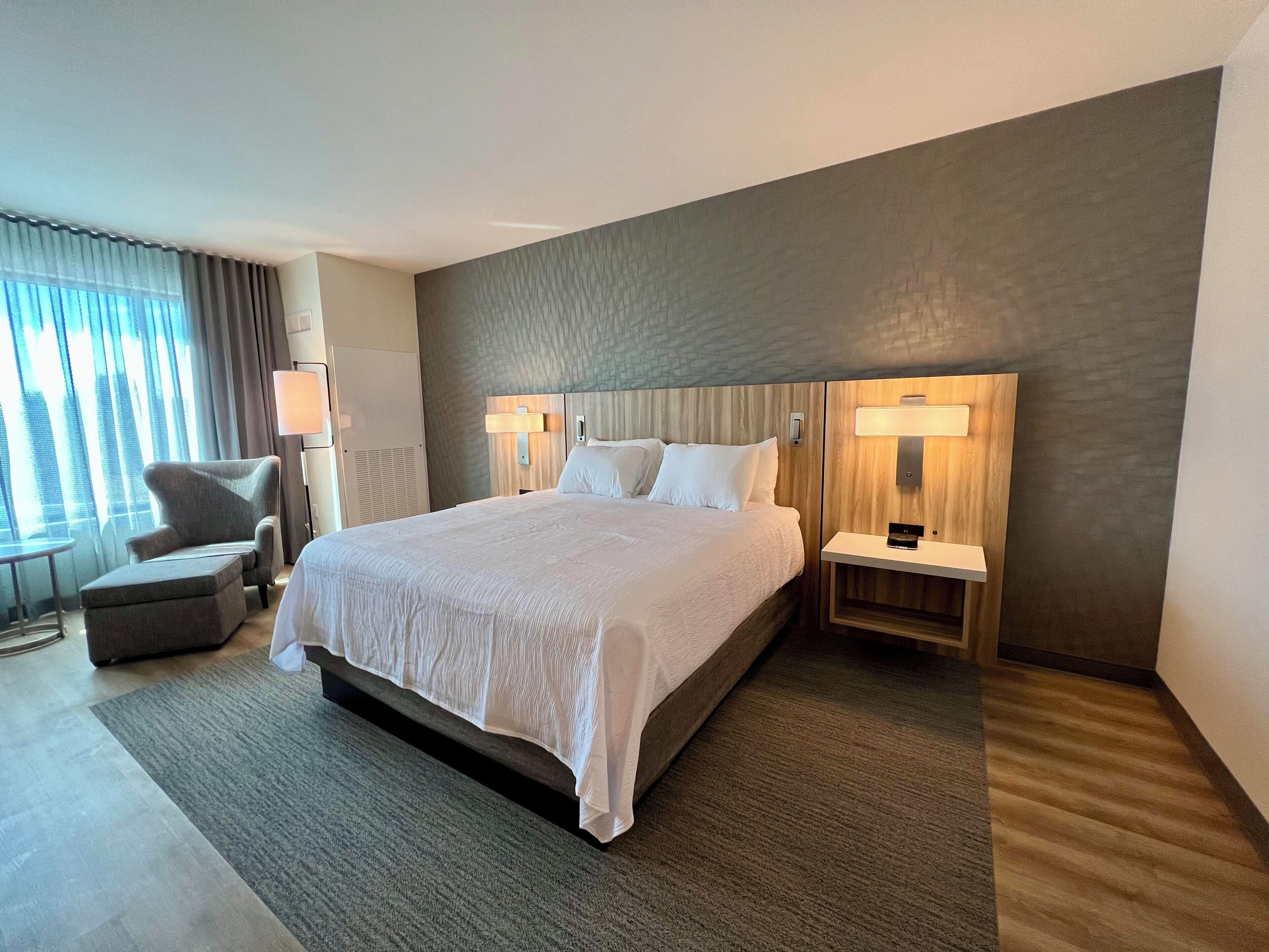 A hotel room with a large bed, two bedside tables with lamps, a chair, and a view of the outdoors through the window.