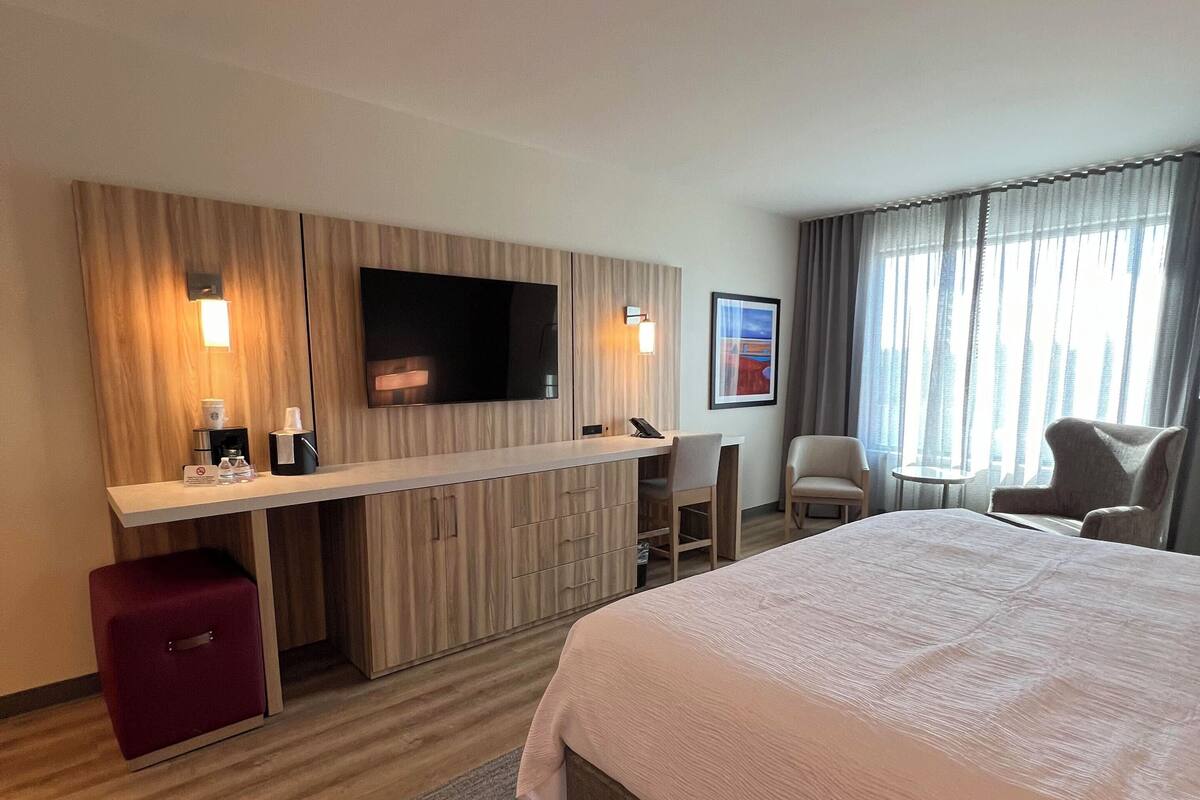 Una habitación de hotel moderna con una cama grande, un escritorio con televisor de pantalla plana, un mueble de almacenamiento rojo y dos sillas.