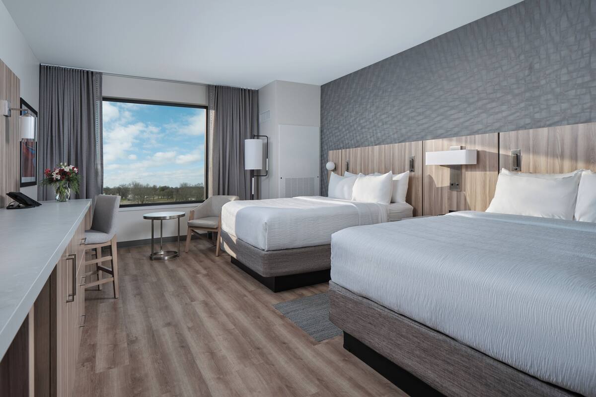 Una habitación de hotel moderna con dos camas, un escritorio, una silla y un amplio ventanal con vistas al exterior.