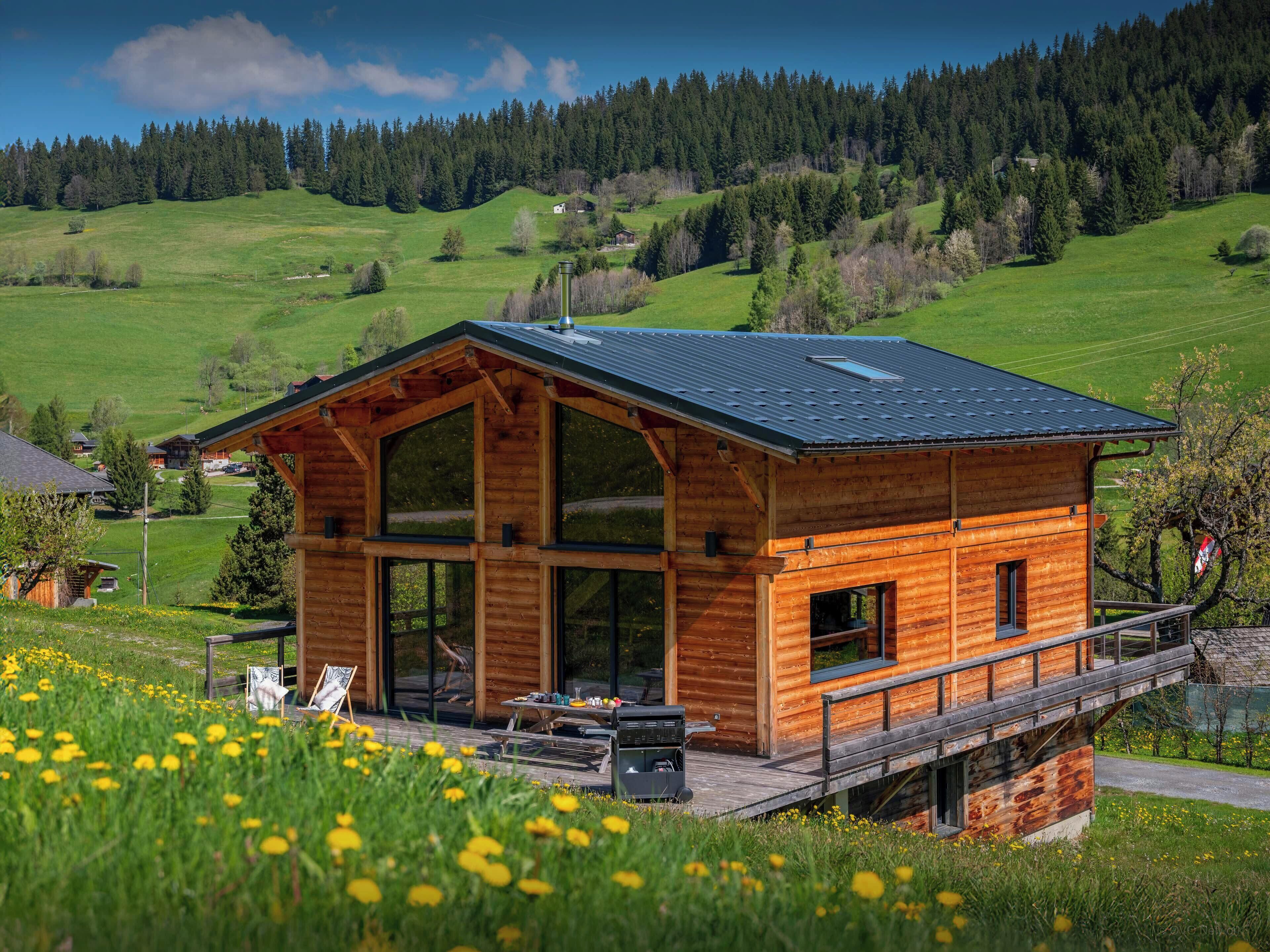 Una cabaña de madera con terraza, ventanas grandes y vistas a un valle verde.