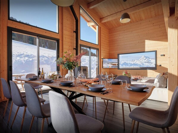 A dining area with a wooden table set for six, large windows offering a snowy mountain view, and a flat-screen TV mounted on the wall.