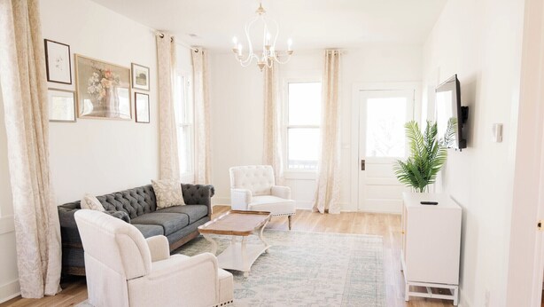 Ein Wohnzimmer mit grauem Sofa, weißen Sesseln, einem Holztisch und einer weißen Kommode mit einer Pflanze.
