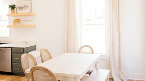Ein Esszimmer mit einem weißen Tisch, vier Holzstühlen, einer Chandelüster und einem Fenster mit Vorhängen.