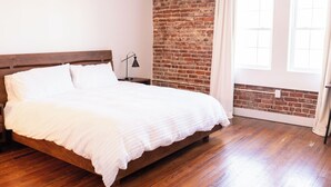 A bedroom with a wooden bed, a brick wall, and a window with curtains. - The Virginian Room @ Main Street Lofts (Victoria)
