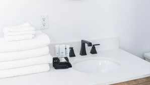 A bathroom with a white sink, black faucet, and a round mirror. There are three clear glass light fixtures above the mirror and a stack of white towels on the counter. - The Virginian Room @ Main Street Lofts (Victoria)