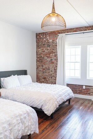 A bedroom with a brick wall, two beds, a hanging light fixture, and a window with curtains. - The Pullman Room @ Main Street Lofts (Victoria)