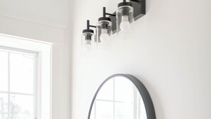 A bathroom with a white vanity, a round mirror, and a window with a view of greenery. - The Darlington Room @ Main Street Lofts (Victoria)