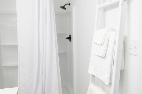 Une salle de bain blanche avec un rideau de douche, un porte-serviettes avec des serviettes pliées et une pomme de douche visible.