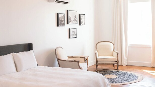 A bedroom with a large bed, a chair, a small table, and a hanging light fixture. - The Victorian Room @ Main Street Lofts (Victoria)