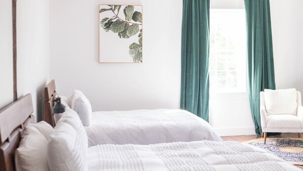 A bedroom with two beds, a wooden headboard, a framed plant, green curtains, and a hanging light fixture. - The Scotsman Room @ Main Street Lofts (Victoria)