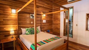 Una cama con dosel y un edredón colorido, una mesita de noche con lámpara, paredes de madera con espejo y un equipo de aire acondicionado.