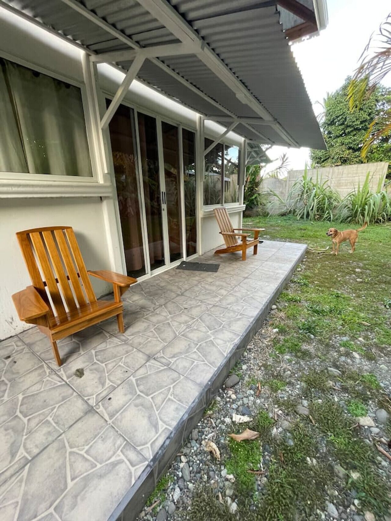 Un patio techado con dos sillas de madera, una mesita y un perro al fondo.