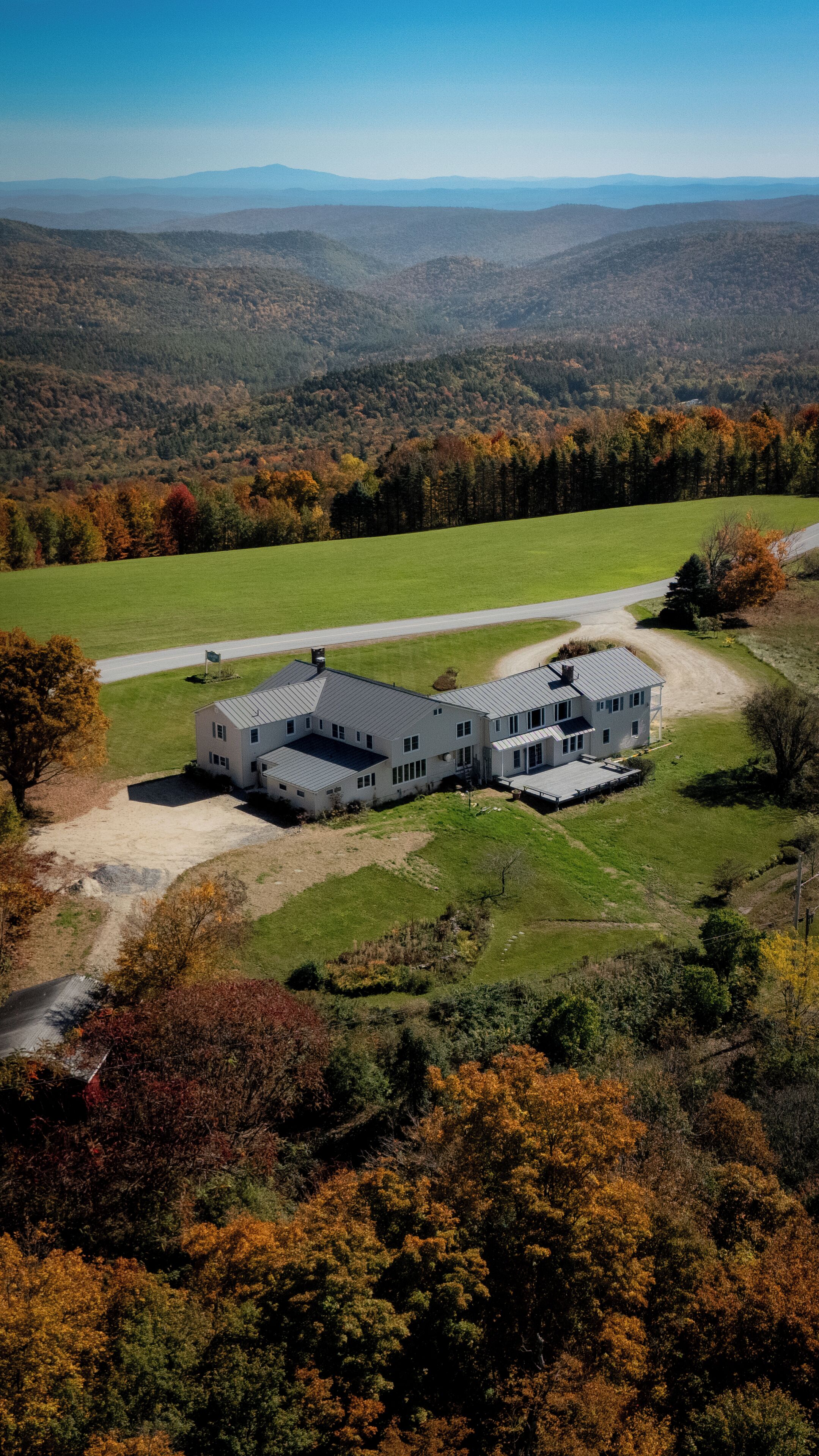 A large, modern two-story house with a spacious driveway and a well-maintained lawn, surrounded by a forested area with autumn foliage.