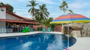 A swimming pool with a rainbow umbrella, green chairs, and a building with a red roof. - Poolside | 2BR Home | Walk to Beach & Dining (Manzanillo)