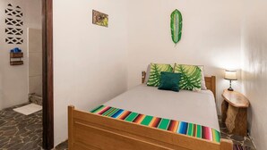 A bed with a colorful striped bedspread, a green leaf decoration on the wall, and a small wooden stool with a lamp beside it. - Serene Poolside Stay | Walk to Beach | Near Dining (Manzanillo)