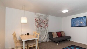 Ein gemütlicher Raum mit Holztisch, zwei Stühlen, einem grauen Sofa, einem blumigen Vorhang, einem gerahmten Bild an der Wand und einem gemusterten Teppich.