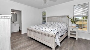 Une chambre à coucher avec un lit en bois, une table de chevet, un ventilateur de plafond, une fenêtre avec des stores et une porte donnant sur une salle de bain.