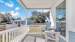 Une véranda couverte avec une balustrade blanche, une table avec des chaises, et une vue sur une maison et des arbres en arrière-plan.