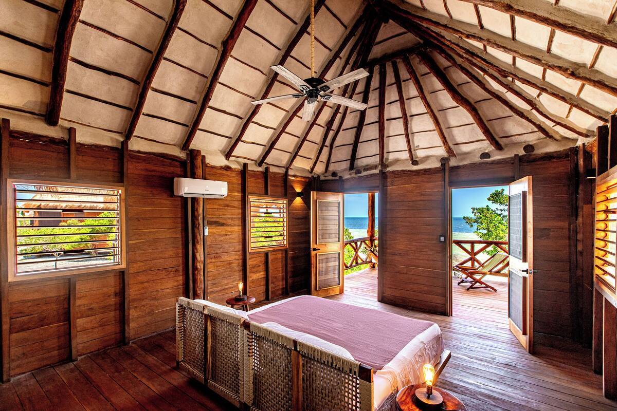 A wooden cabin with a bed, a ceiling fan, and a balcony with a view of the sea.