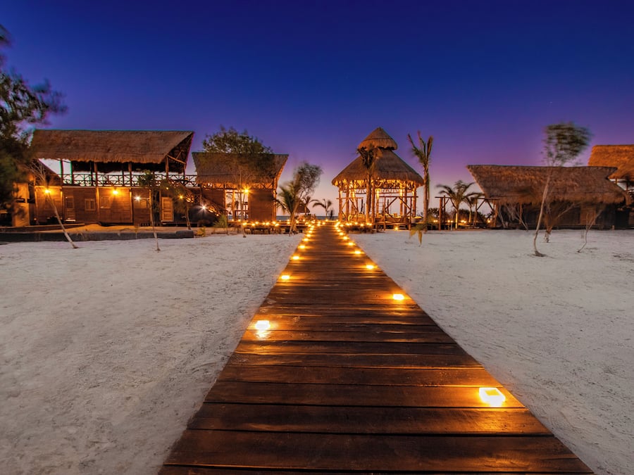 Un sendero de madera que conduce a un complejo turístico frente a la playa con construcciones de techo de paja.