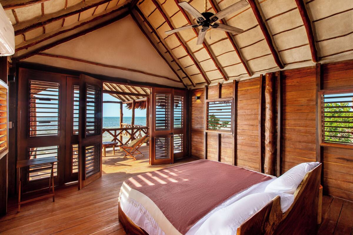 A wooden room with a bed, a ceiling fan, and a view of the sea.