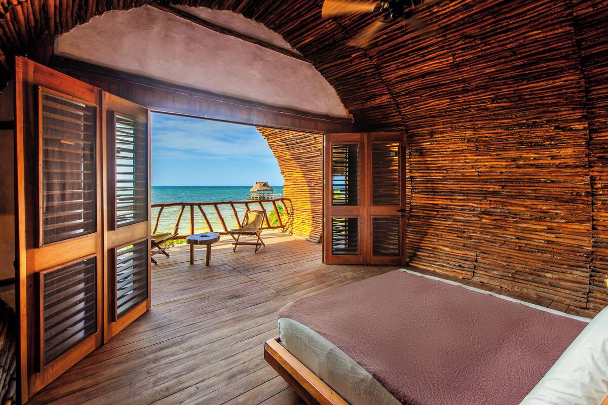 A room with a wooden floor, a bed with a brown blanket, and a view of the ocean through open shutters.