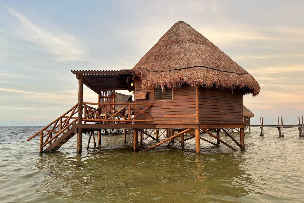 A wooden overwater bungalow with a thatched roof, wooden deck, and a small covered area.