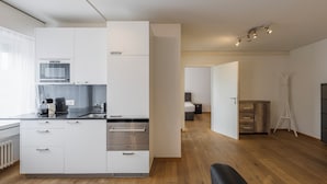 Una cocina moderna con gabinetes blancos, horno empotrado y microondas. Hay una zona de comedor con una silla negra y una mesa pequeña. El piso es de madera y el techo es blanco.