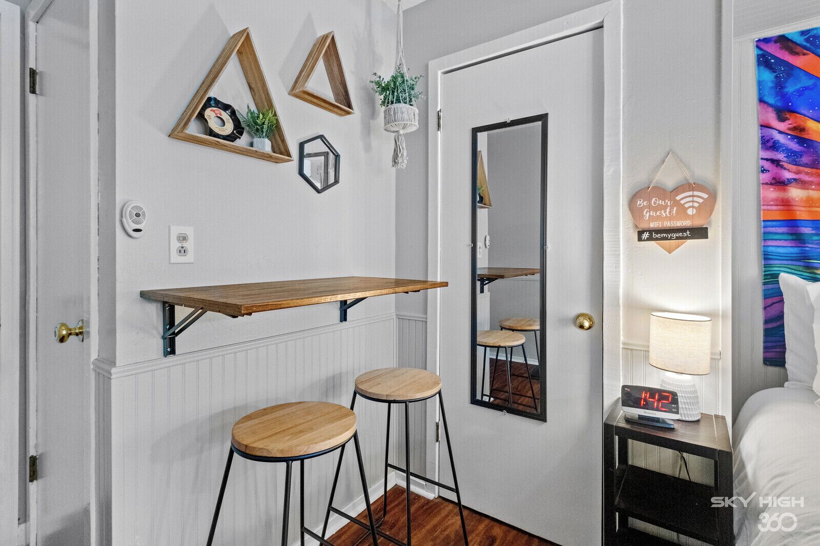 A small room with a wooden bar stool, a mirror, and a wall-mounted shelf with decorative items.