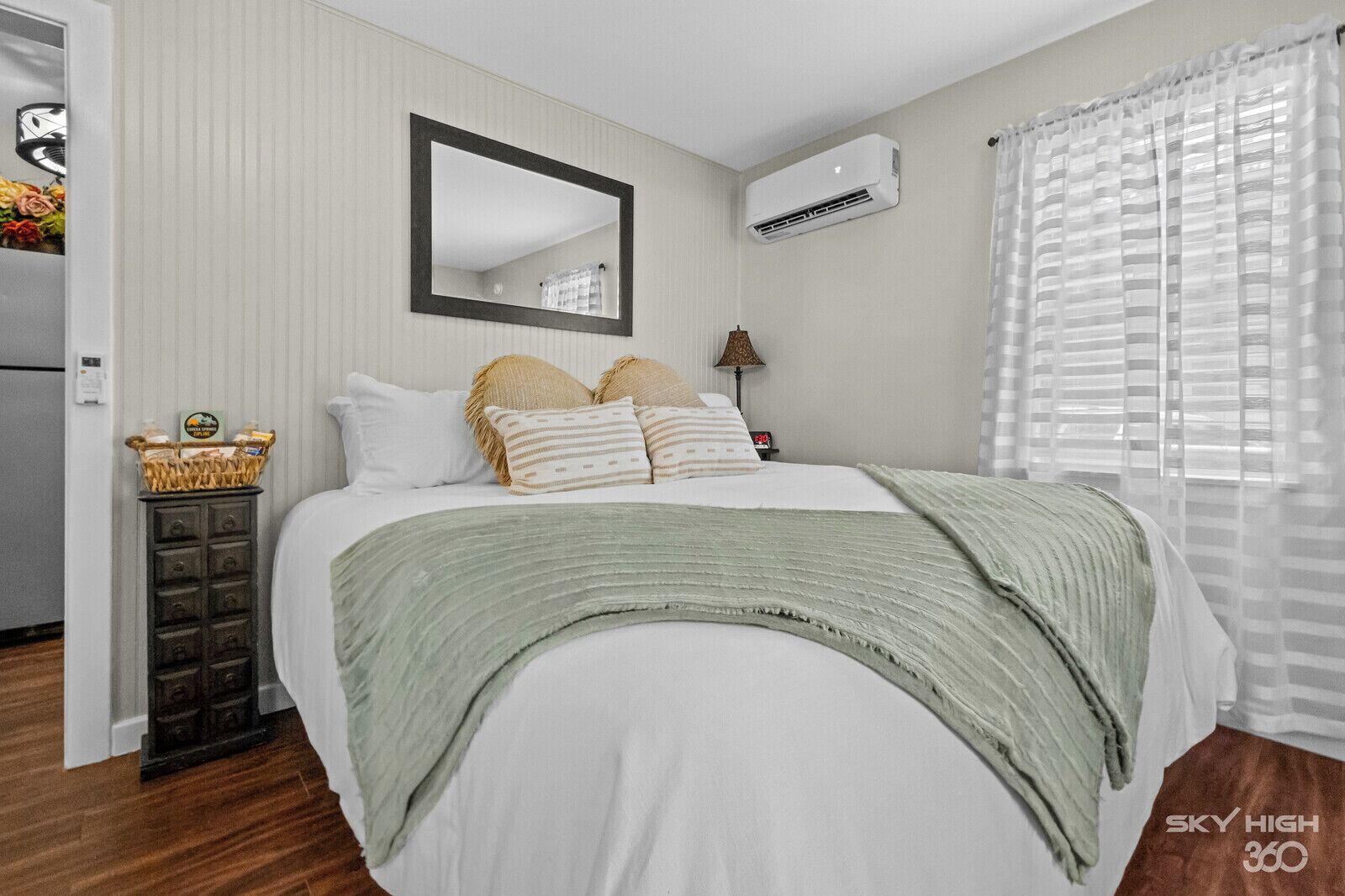 A bedroom with a bed, a mirror, an air conditioner, a lamp, and a window with blinds.