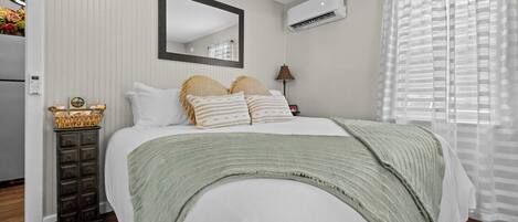 A bedroom with a bed, a mirror, an air conditioner, a lamp, and a window with blinds.