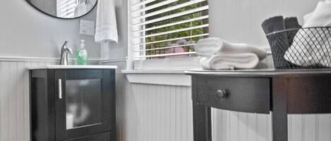 A bathroom with a wooden vanity, a round mirror, a window with blinds, and a towel rack.