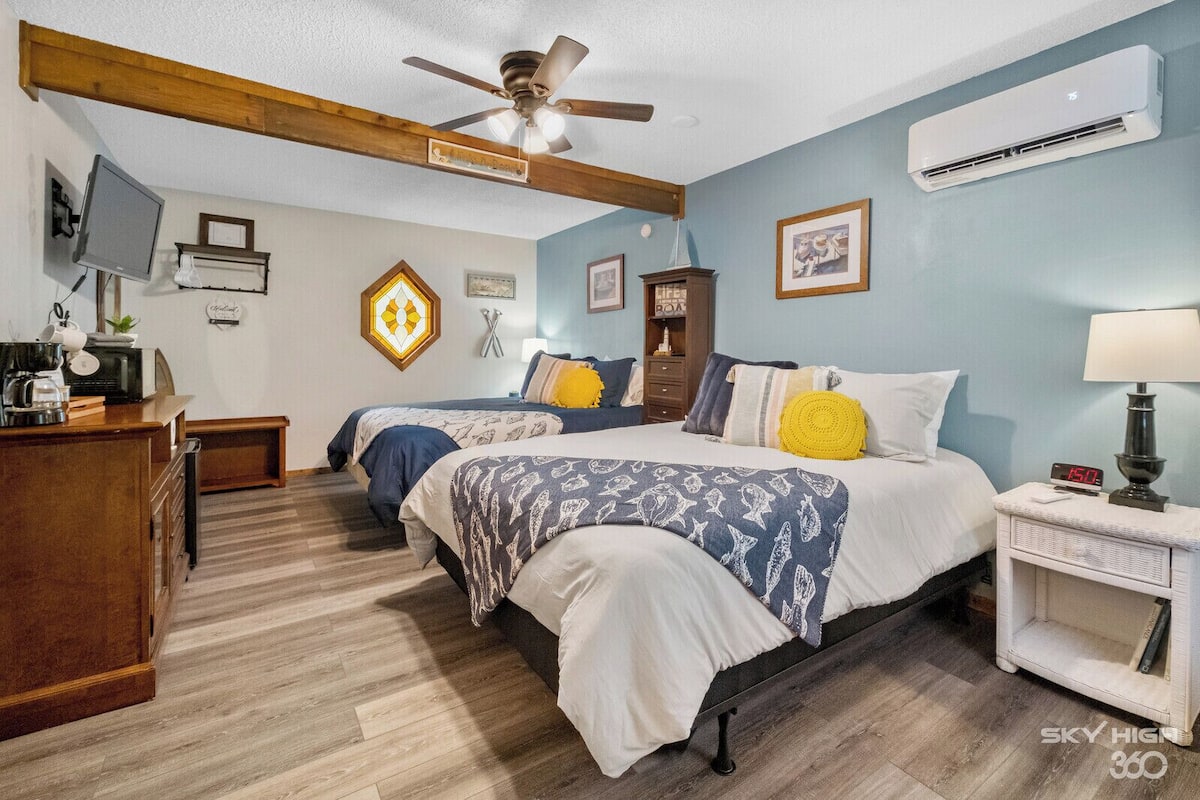 A hotel room with two beds, a wooden ceiling, a ceiling fan, a TV, a coffee maker, a small table, and a bedside lamp.