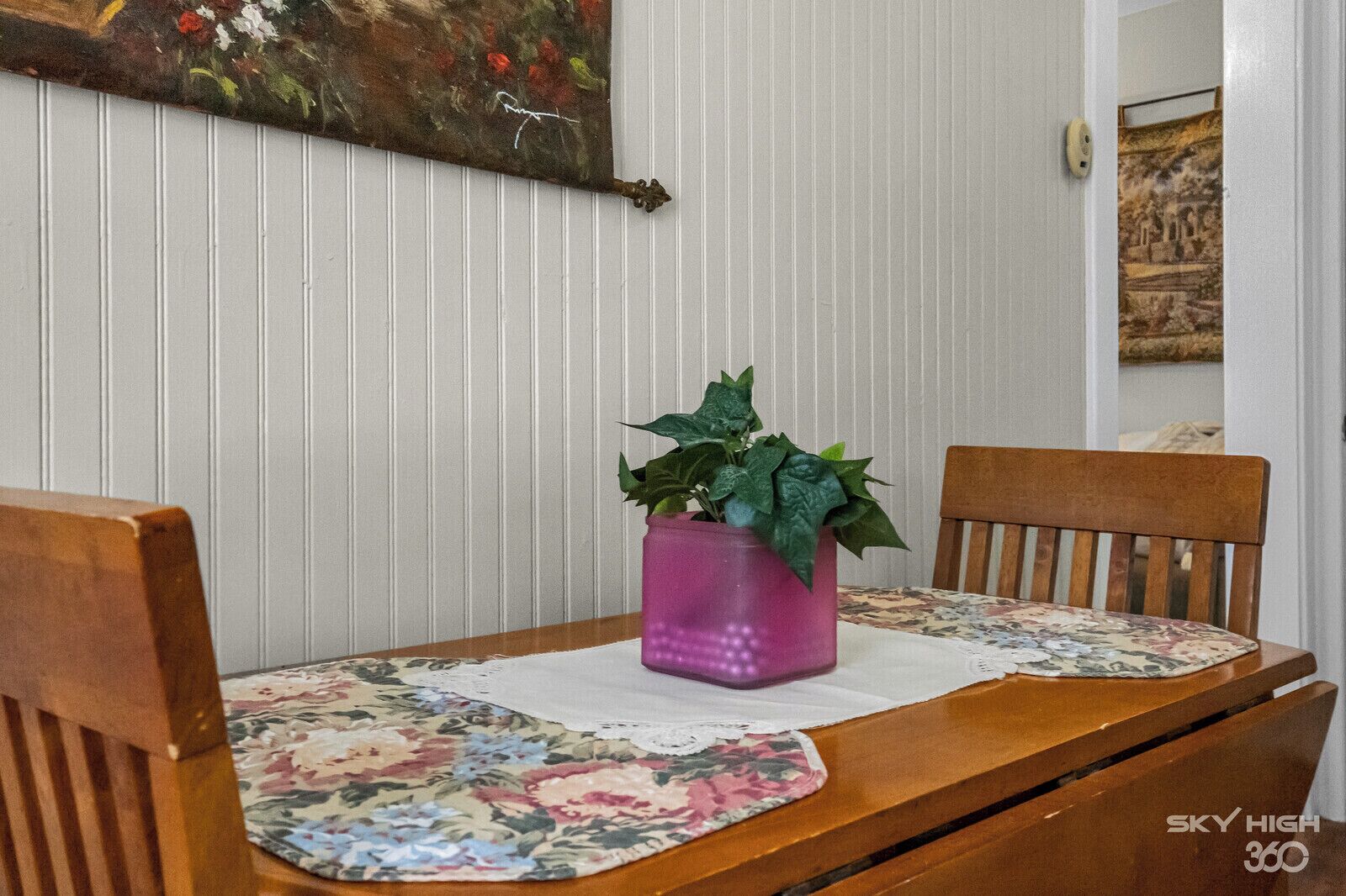 A dining table with a floral placemat, two wooden chairs, and a potted plant in a pink vase.
