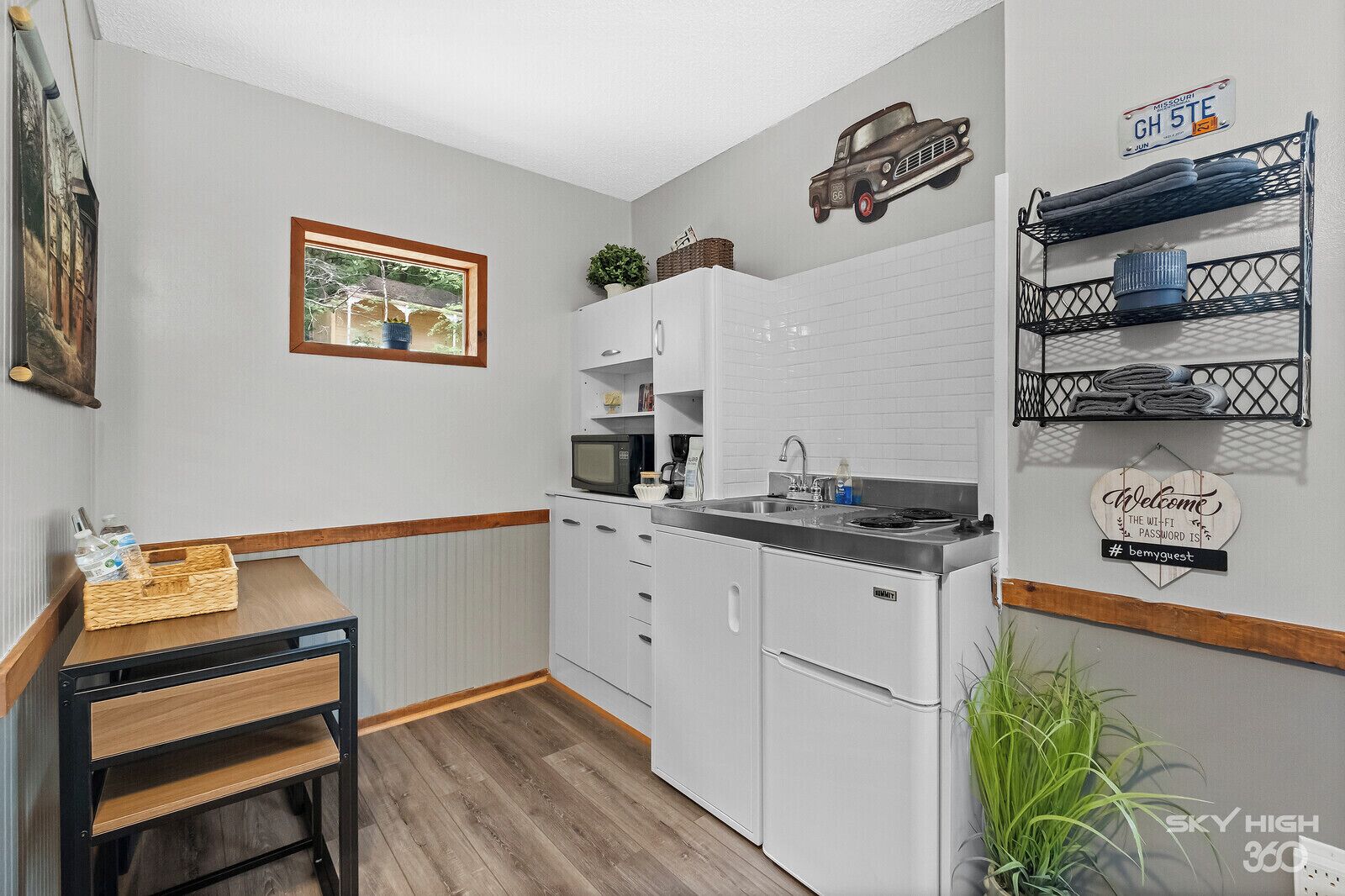 A compact kitchen with white cabinets, a microwave, and a sink. There's a small table with a basket, a framed picture, and a welcome sign with a hashtag.